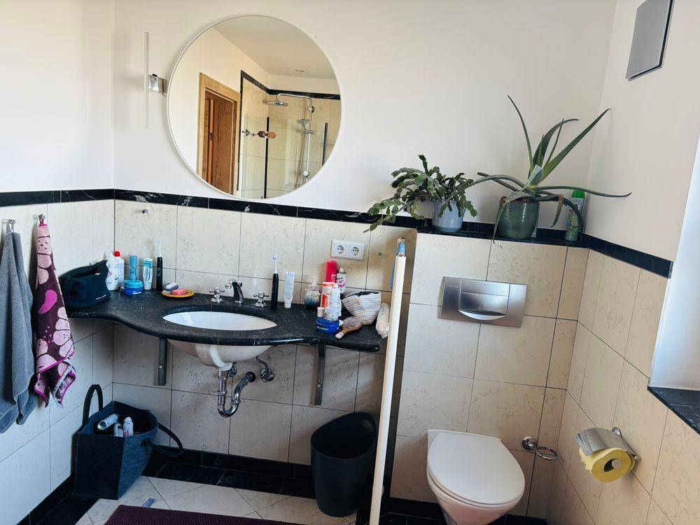 Badezimmer mit Fliesen, Einzelwaschbecken, Toilette und großem Rundspiegel.