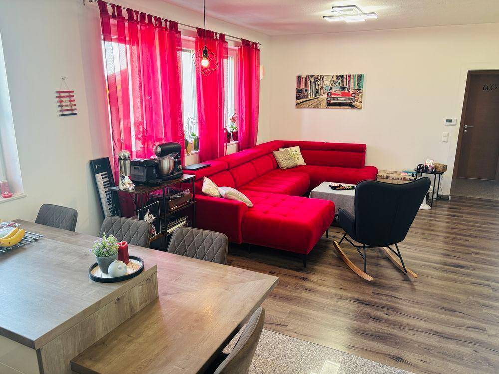 Gemütliches Wohnzimmer mit rotem Ecksofa, Schaukelstuhl und Essbereich, beleuchtet durch große Fenster.