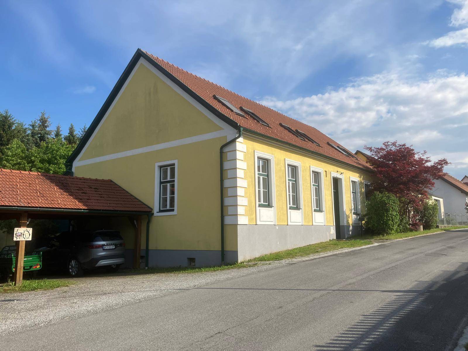 Traditionelles gelbes Wohnhaus mit rotem Ziegeldach und Carport entlang einer ruhigen Straße.