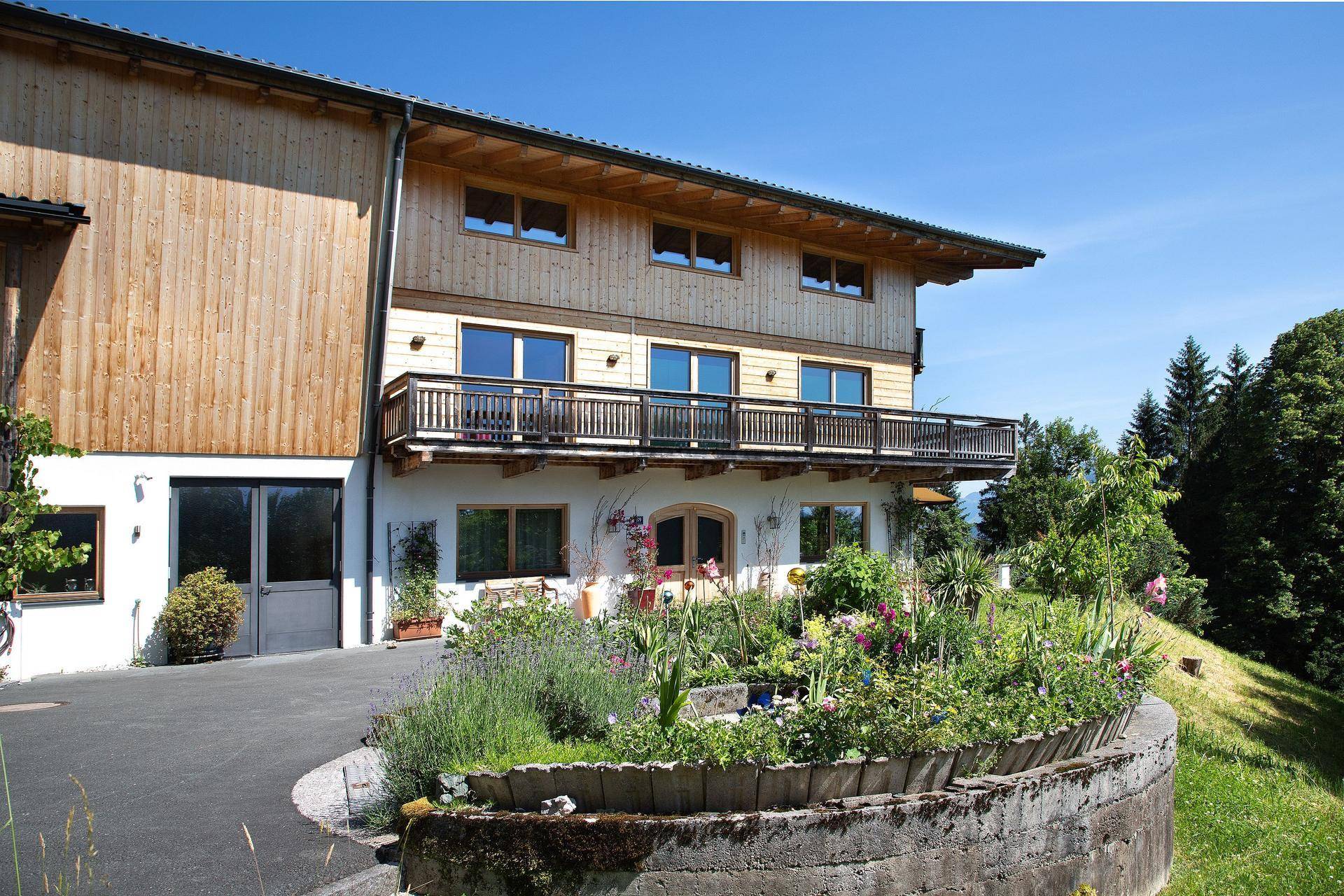 Seitenansicht des Chalets mit Holzelementen, Balkon, Auffahrt und einem liebevoll bepflanzten Blumenbeet.