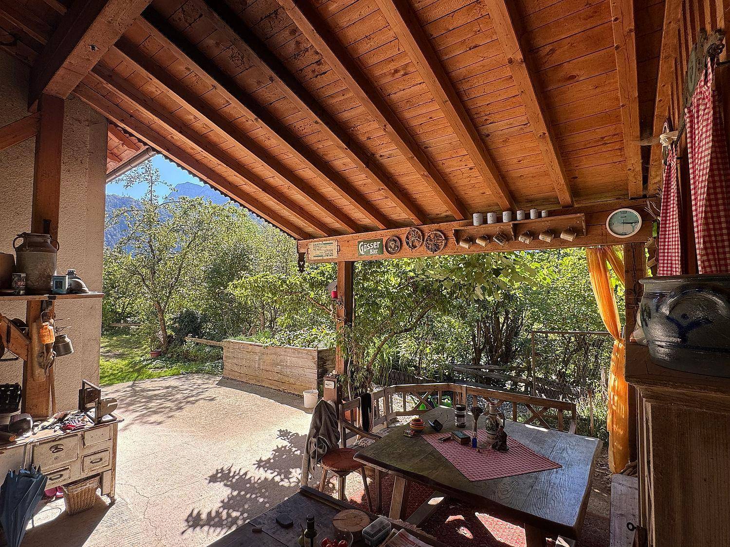 Gemütliche überdachte Terrasse mit Holztisch und Bänken, umgeben von viel Grün.