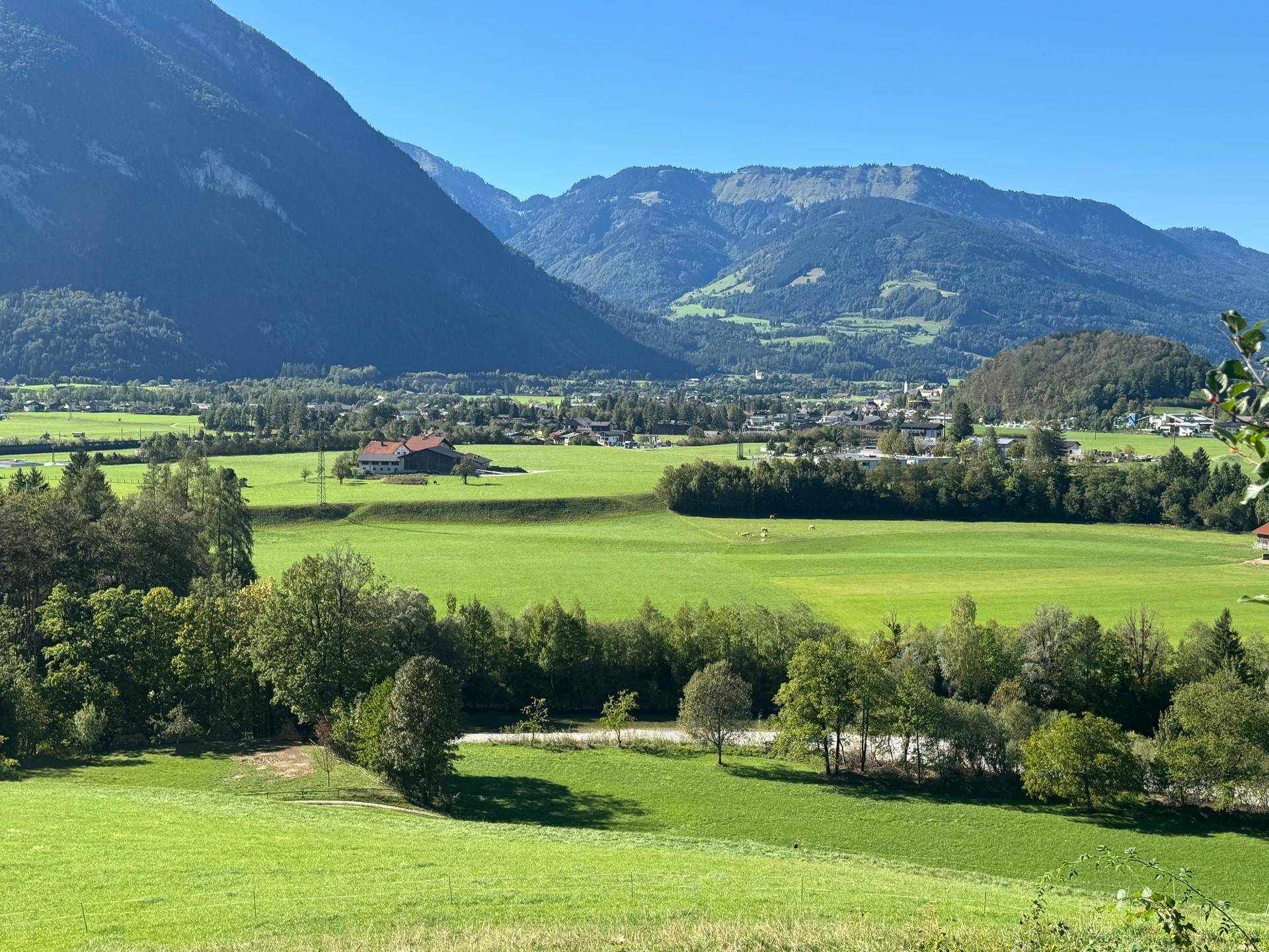 Panoramablick über ein weites grünes Tal mit Bergen und einer Siedlung in der Ferne.