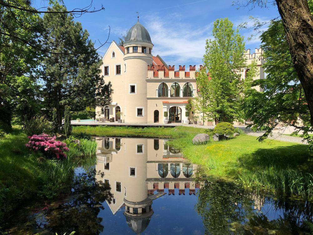 Majestätisches Anwesen mit Zinnen und Turm, umgeben von üppigem Grün und einem spiegelnden Teich.