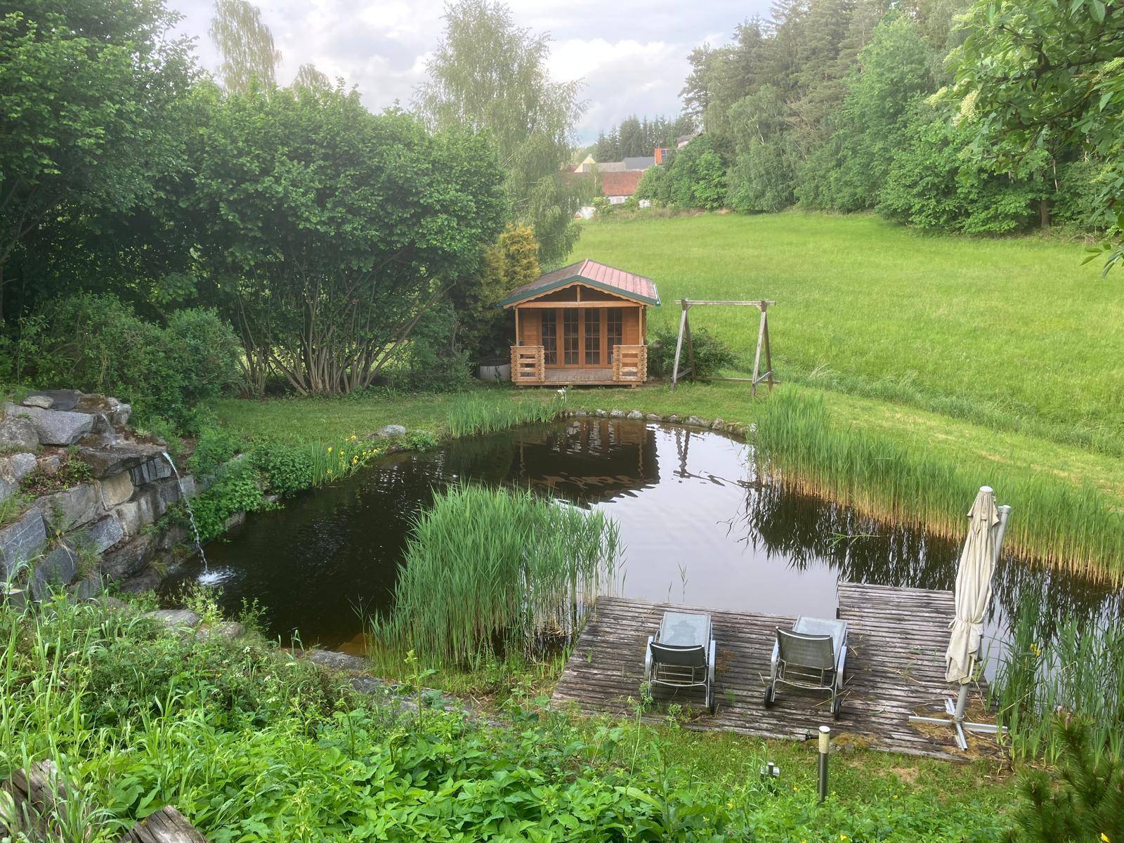 Weitläufiger Garten mit Naturteich, Holzsteg, Gartenhaus und Schaukel in grüner Umgebung.