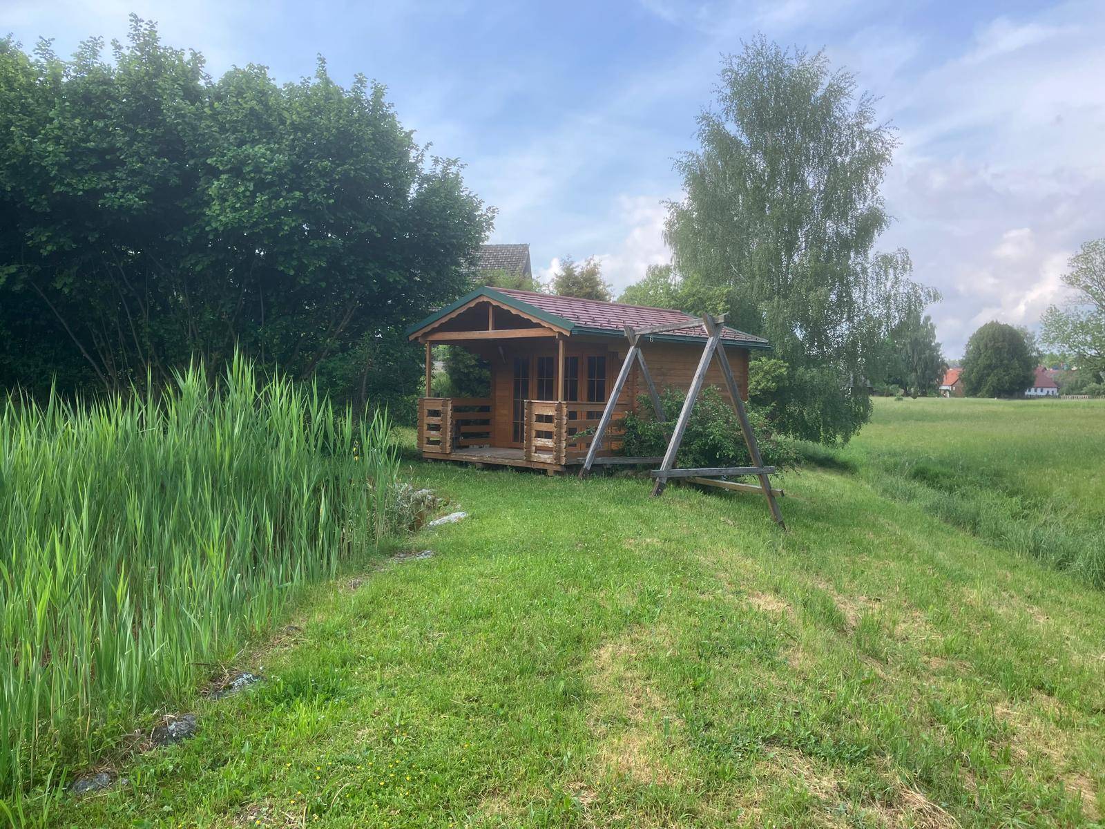 Rustikales Gartenhaus aus Holz mit Veranda und Schaukel in einer weitläufigen Wiesenlandschaft.