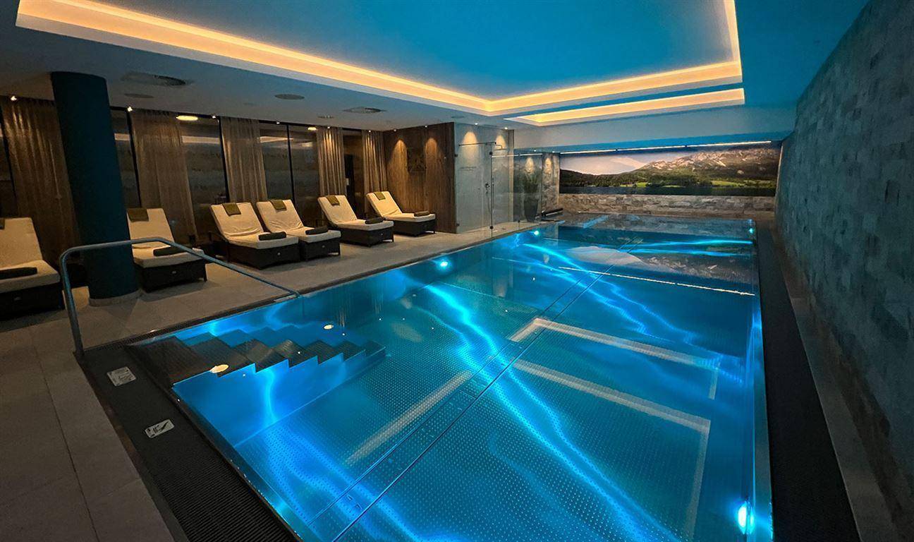 Beleuchteter Innenpool im modernen Wellnessbereich mit Liegeflächen und Bergblick-Wandbild.