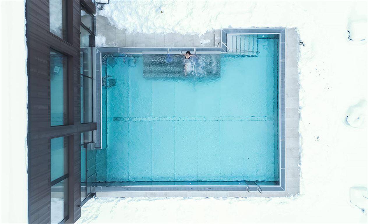 Luftaufnahme eines beheizten Außenpools, umgeben von Schnee und moderner Architektur.