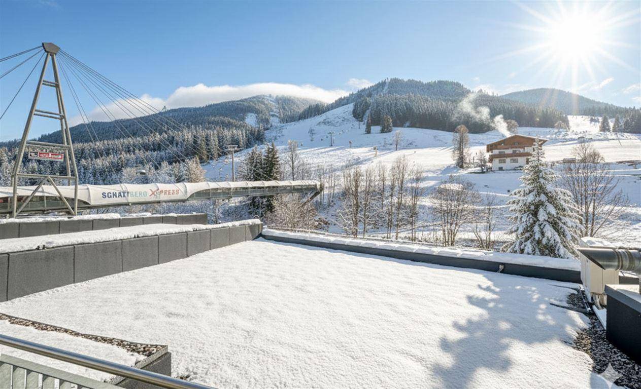 Schneebedeckte Dachfläche mit Blick auf die verschneiten Berge und eine Skiliftanlage.