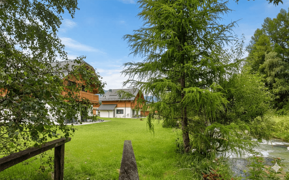Ferienhäuser in idyllischer grüner Umgebung mit Bäumen und einem fließenden Gewässer.