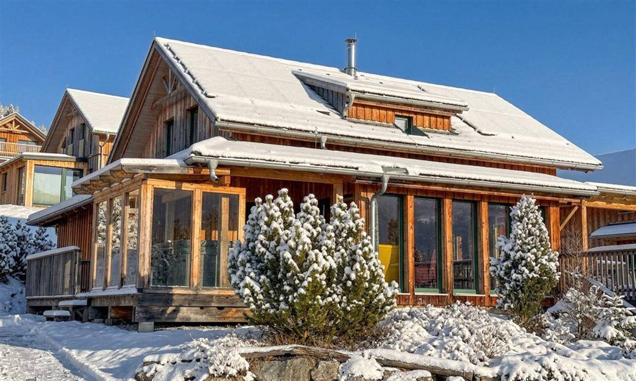 Idyllisches Holzhaus mit Solarpaneelen auf dem schneebedeckten Dach in winterlicher Landschaft.