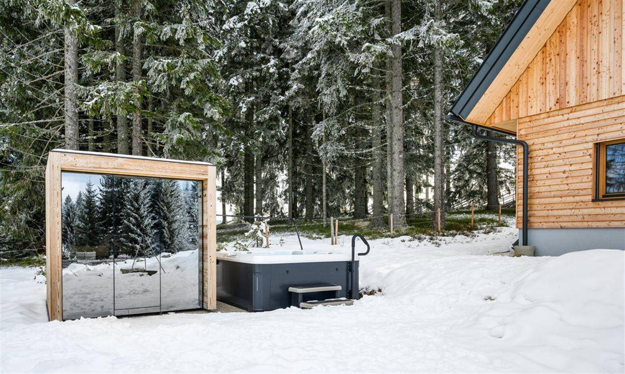 Verschneite Gartenanlage mit Whirlpool und Sauna vor einem dichten Waldhintergrund.
