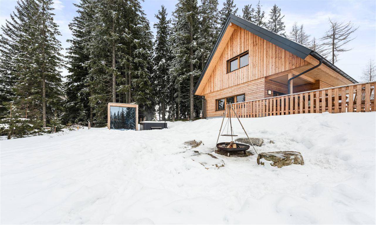 Verschneite Außenanlage mit Feuerstelle, Whirlpool und Sauna vor dem Holzhaus.