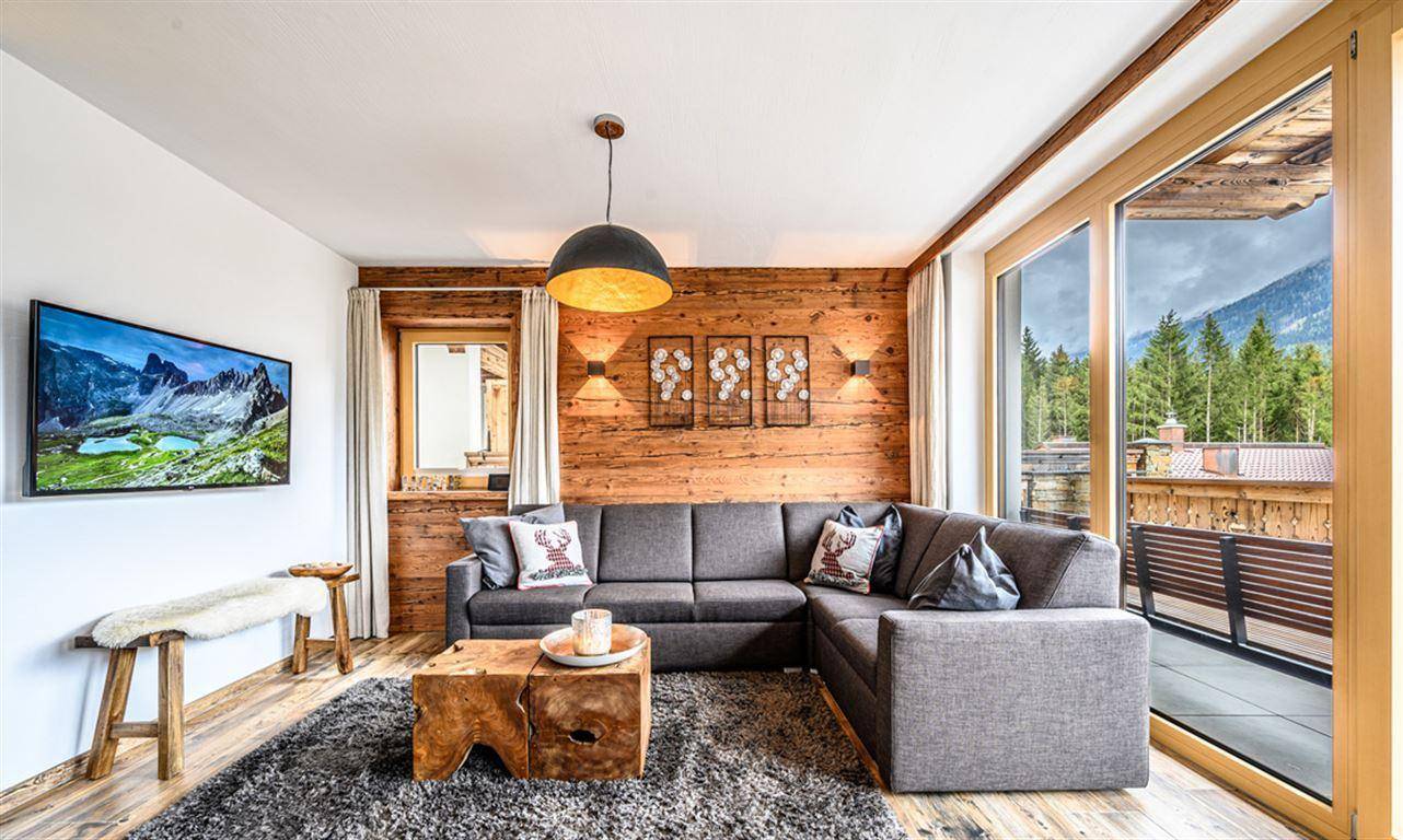 Lichtdurchflutetes Wohnzimmer mit großen Fenstern, Holzwand und Zugang zu einem Balkon mit Bergblick.
