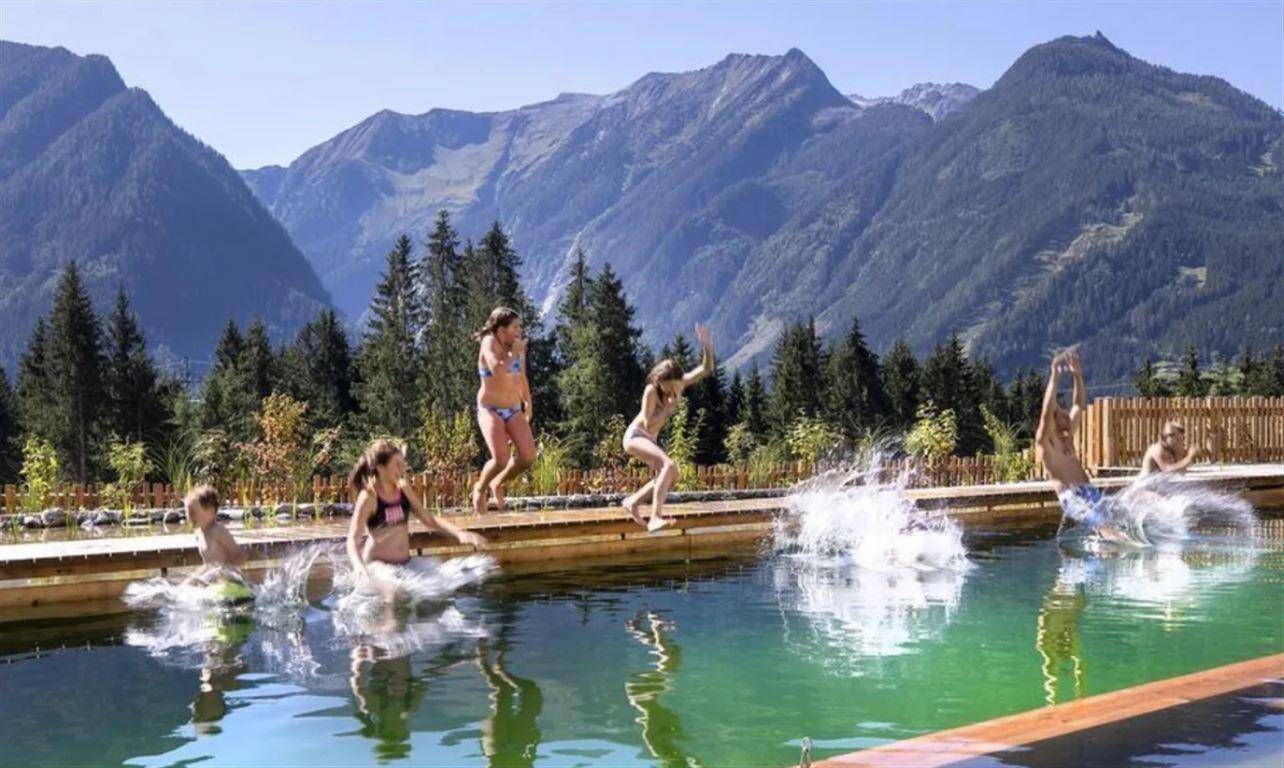 Natürlicher Schwimmteich mit Kindern beim Spielen, umgeben von Bergen und Wald.