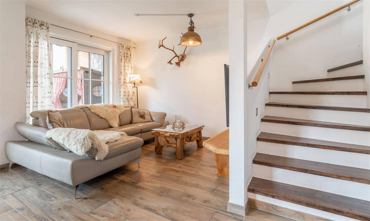 Heller Wohnbereich mit beigem Sofa, Fliesenboden und Holztreppe zum oberen Stockwerk.