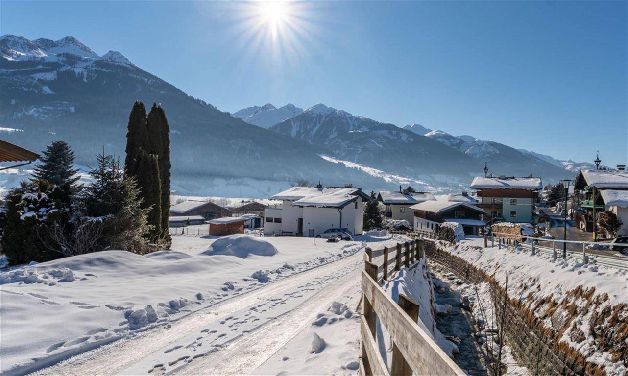 Idyllische Winterlandschaft mit verschneiter Straße, Häusern und majestätischem Alpenpanorama.