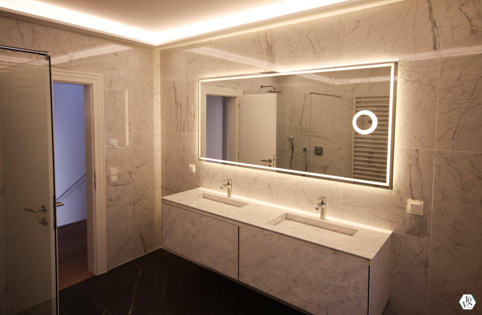 Modernes Badezimmer mit Doppelwaschbecken, beleuchtetem Spiegel und eleganten Marmorfliesen an Wänden und Boden.