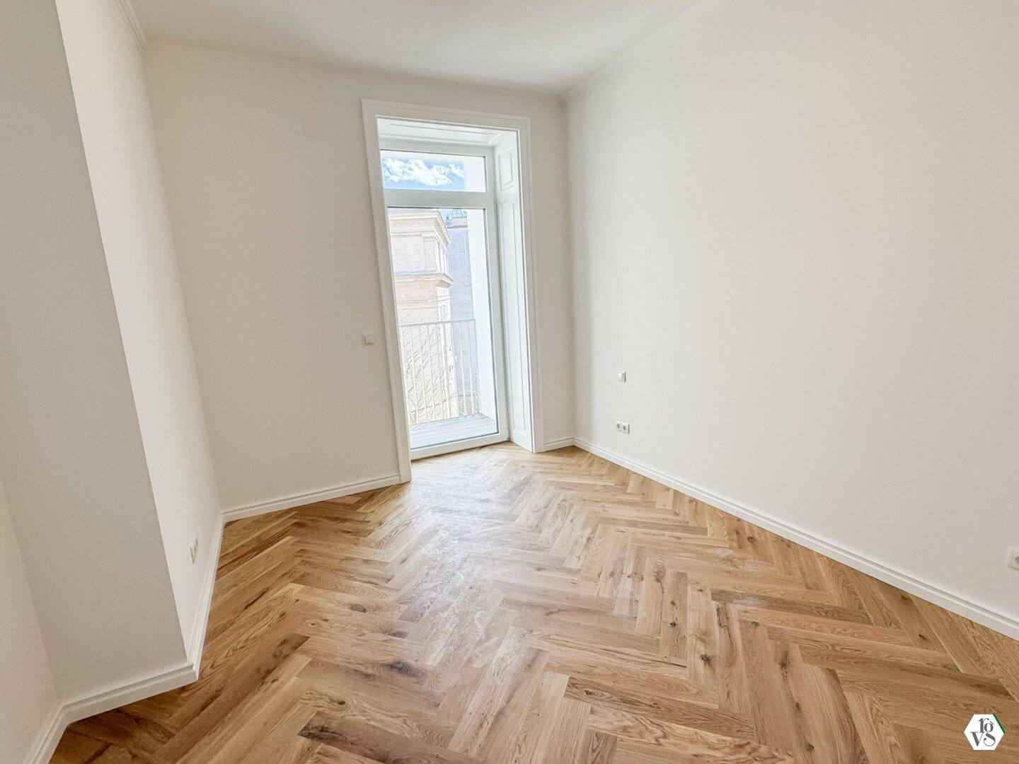 Heller Raum mit Fischgrätparkett und Zugang zu einem französischen Balkon, bietet viel Tageslicht.