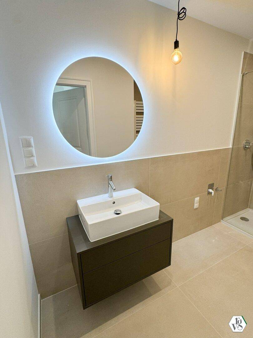 Modernes Badezimmer mit schwebendem Waschtisch, beleuchtetem Spiegel und großformatigen Fliesen.