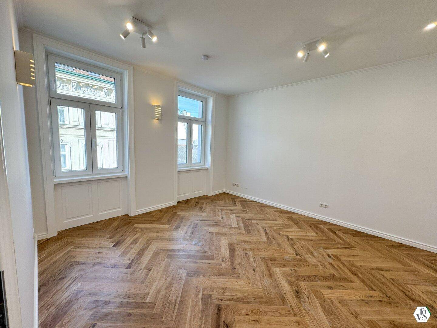 Geräumiges, lichtdurchflutetes Zimmer mit elegantem Fischgrätparkett und moderner Deckenbeleuchtung.
