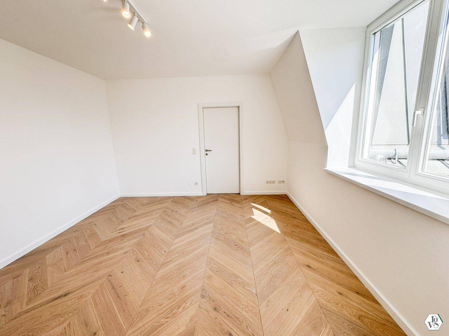 Lichtdurchflutetes Zimmer mit Fischgrätparkett und schrägen Wänden im Dachgeschoss.