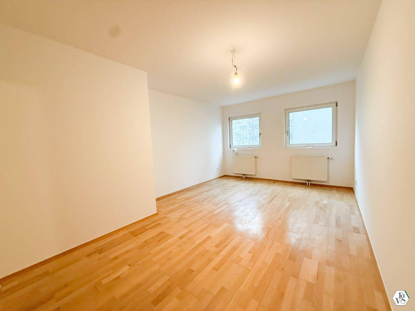 Leeres Zimmer mit Parkettboden und zwei Fenstern.