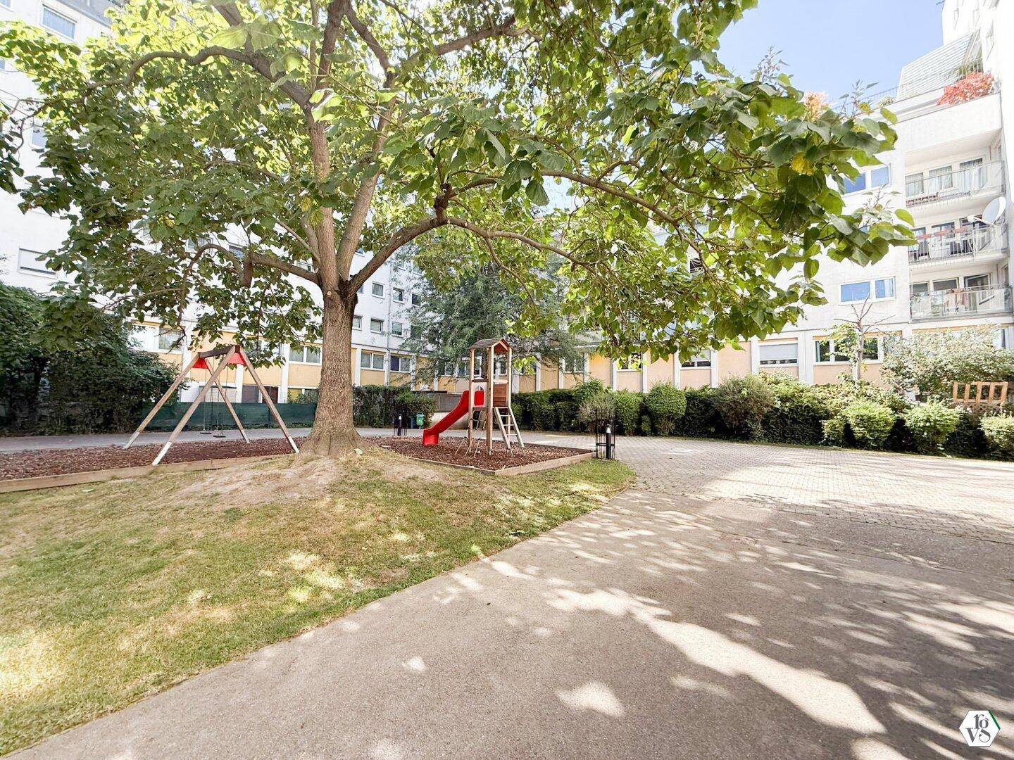 Grüner Innenhof mit Spielplatz, großen Bäumen und Sitzgelegenheiten.