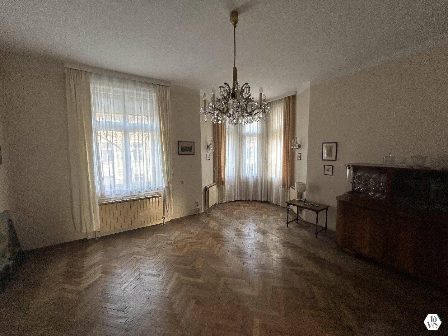 Geräumiges Wohnzimmer mit Fischgrätparkett, großen Fenstern und einem eleganten Kronleuchter.