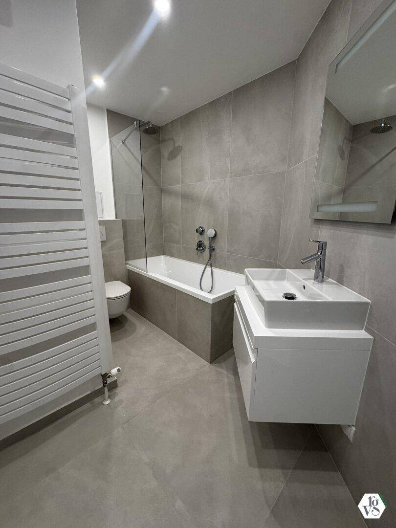 Modernes Badezimmer mit grauen Fliesen, Badewanne mit Duschfunktion und beheiztem Handtuchhalter.