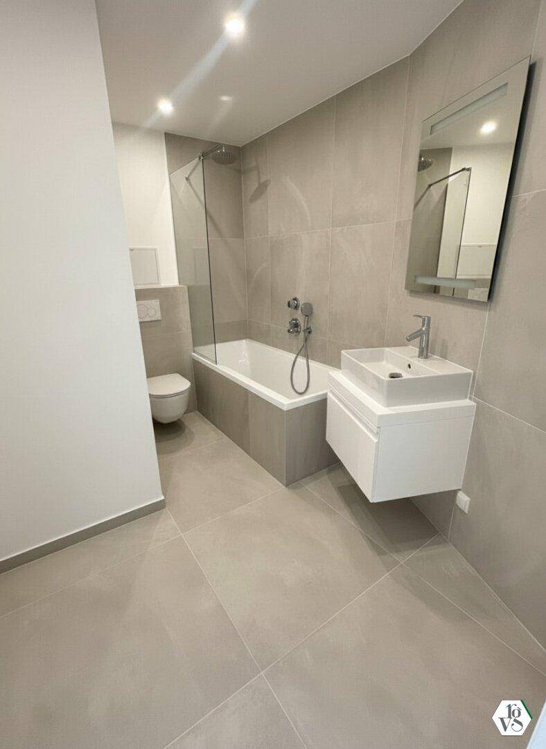 Modernes Badezimmer mit hellgrauen Fliesen, Badewanne mit Duschfunktion und wandhängendem WC.
