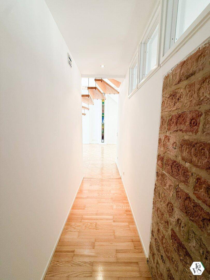 Schmaler Flur mit Holzboden und einer Wand mit freiliegenden Ziegeln, die zu einem weiteren Raum führt.