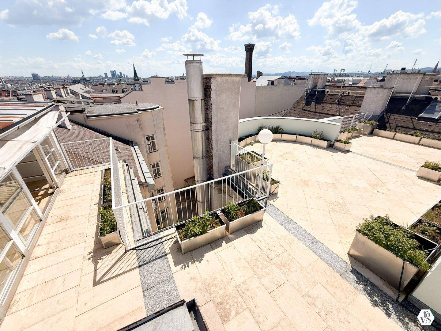Geräumige Dachterrasse mit Stadtblick, umgeben von niedrigen Mauern und Pflanzen in Metallkübeln.
