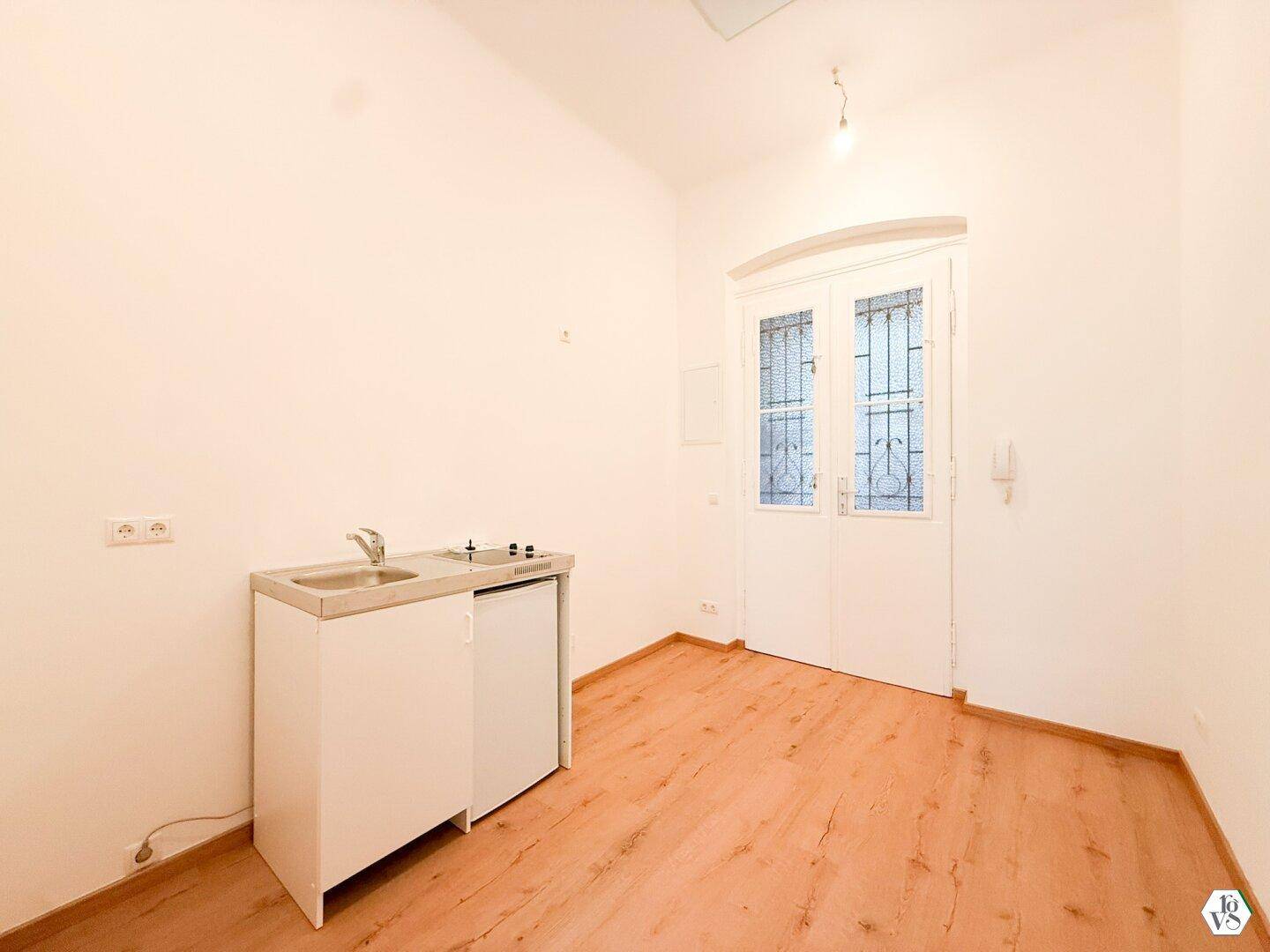 Kompakte Küchenzeile mit Spüle und Kochfeld in einem hellen Raum mit Holzboden und Doppeltür.