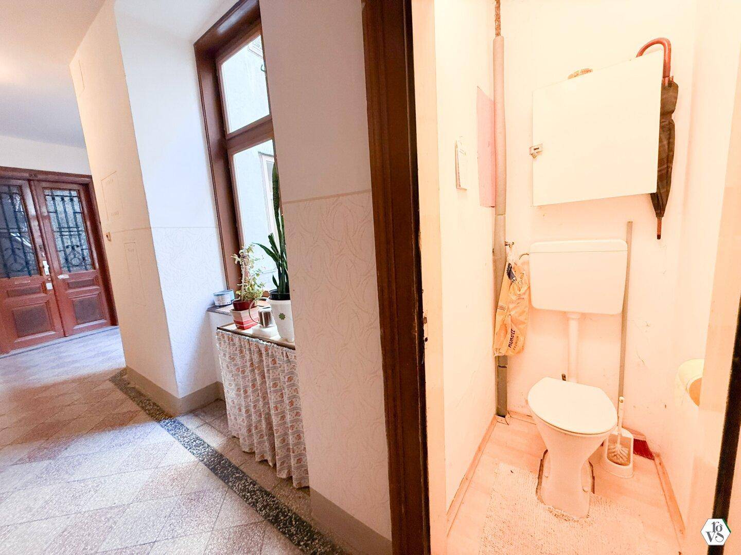 Schmale Toilette mit Fenster und einfacher Ausstattung, angrenzend an einen Flur mit Terrazzoboden.