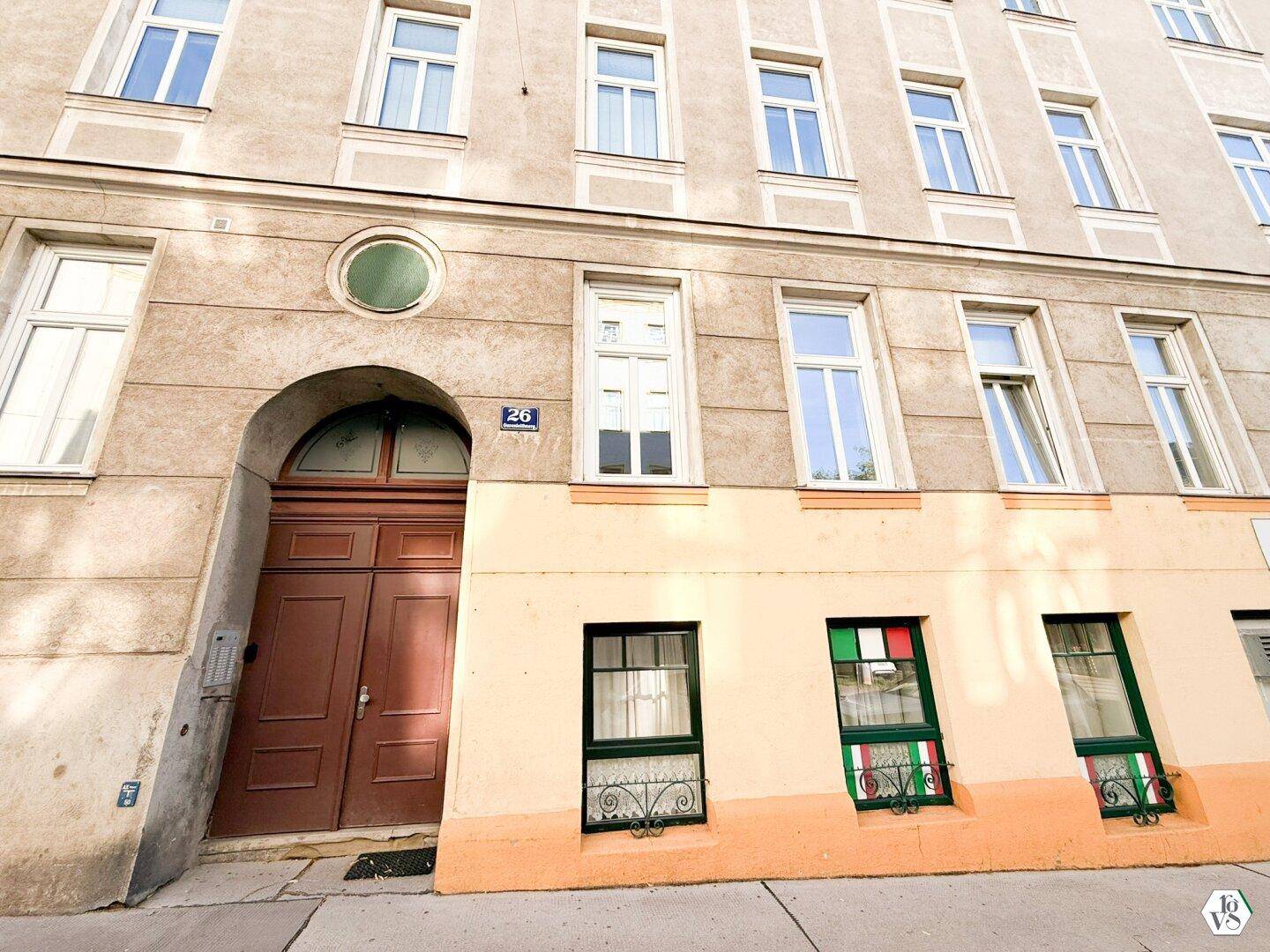 Historische Gebäudefassade mit bogenförmigem Eingang und vielen Fenstern, im Erdgeschoss mit Geschäften.