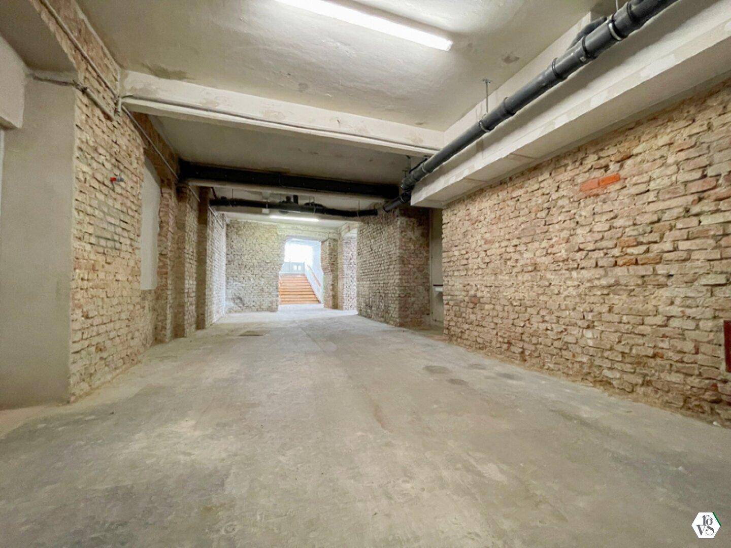 Langer Flur mit unverputzten Ziegelwänden, Betonboden und sichtbaren Rohren an der Decke, führt zu einer Treppe.