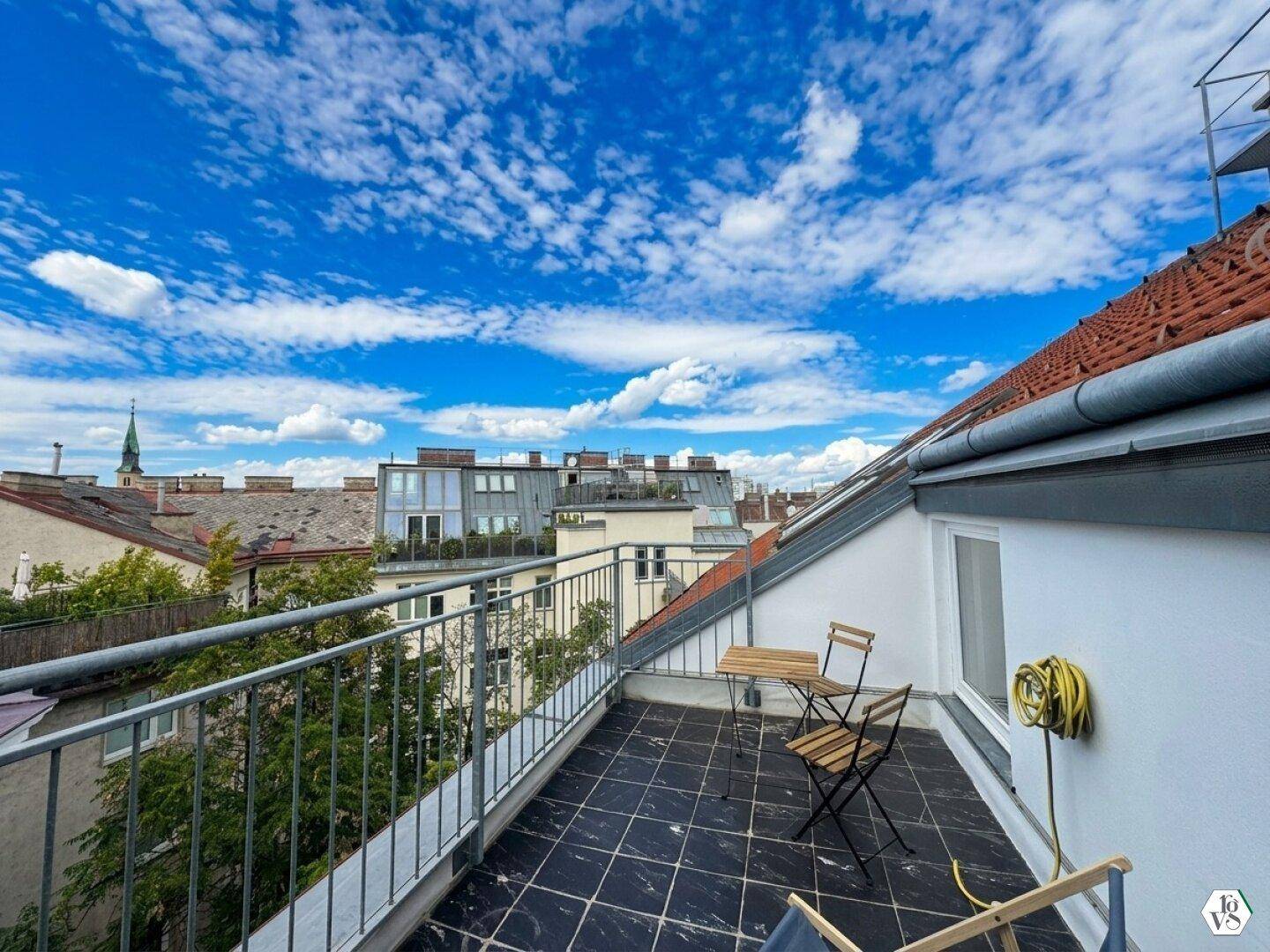Schmaler Balkon mit Fliesenboden, Sitzgelegenheiten und weitem Blick über die Dächer der Stadt.