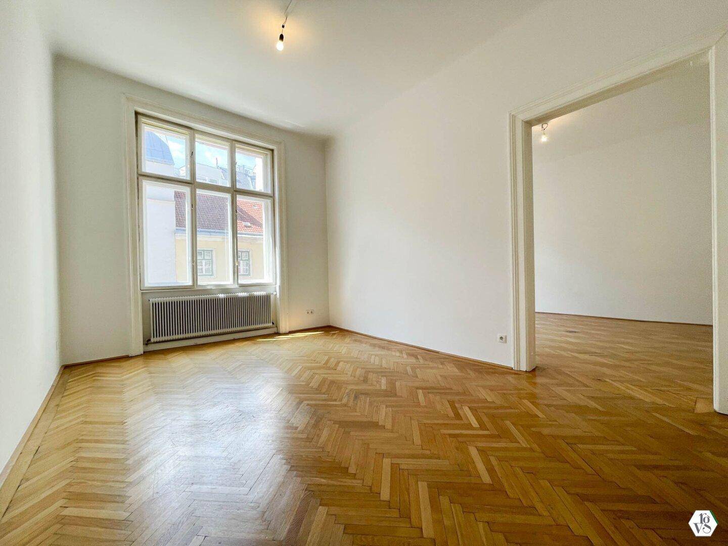 Geräumiges Wohnzimmer mit klassischem Fischgrätparkett und großem Fenster, das viel Licht hereinlässt.