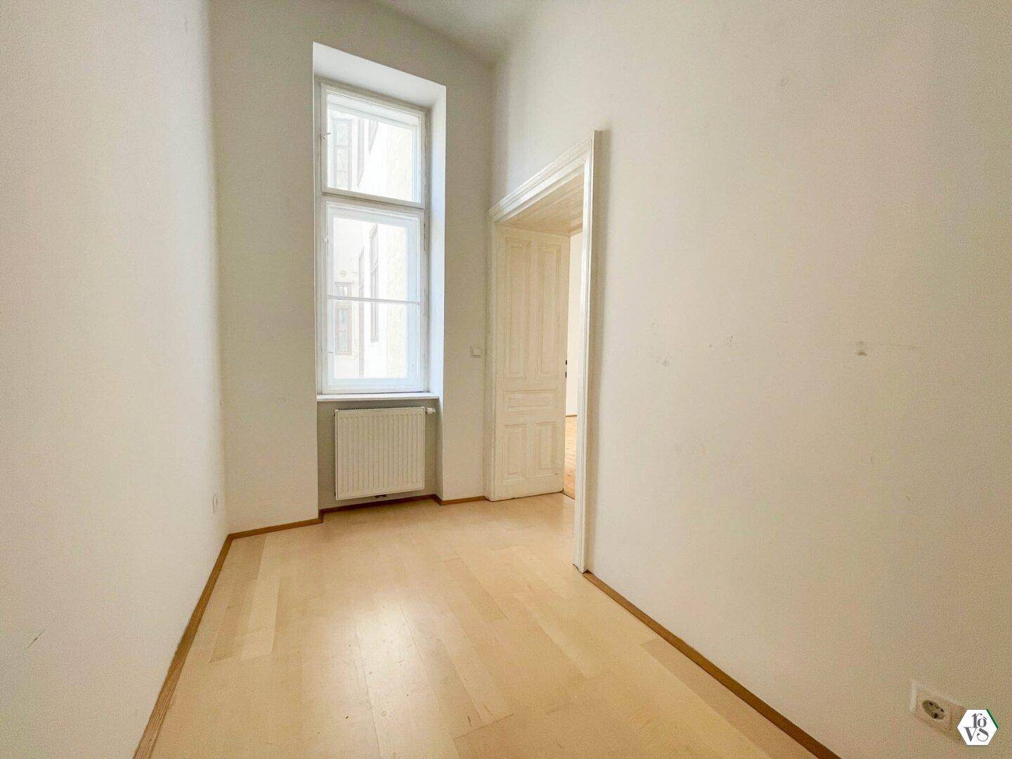 Schmaler Raum mit hellem Holzboden und einem hohen Fenster, das für ausreichend Tageslicht sorgt.