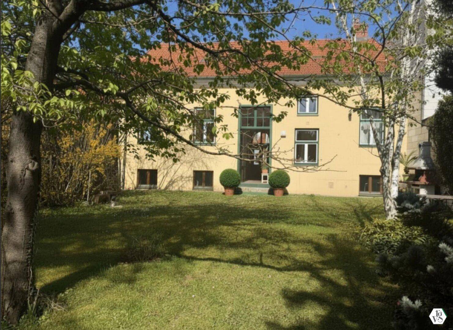 Historisches Gebäude mit gelber Fassade, umgeben von einem gepflegten Garten und altem Baumbestand.