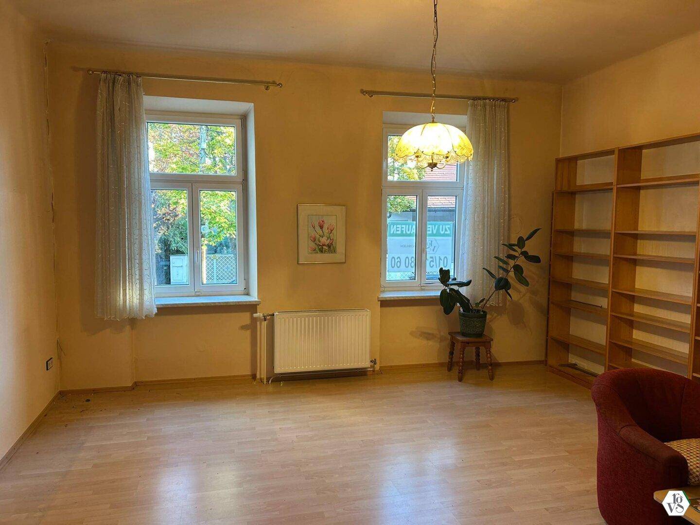 Helles Wohnzimmer mit Parkettboden, zwei großen Fenstern, Heizkörper und einem Bücherregal.