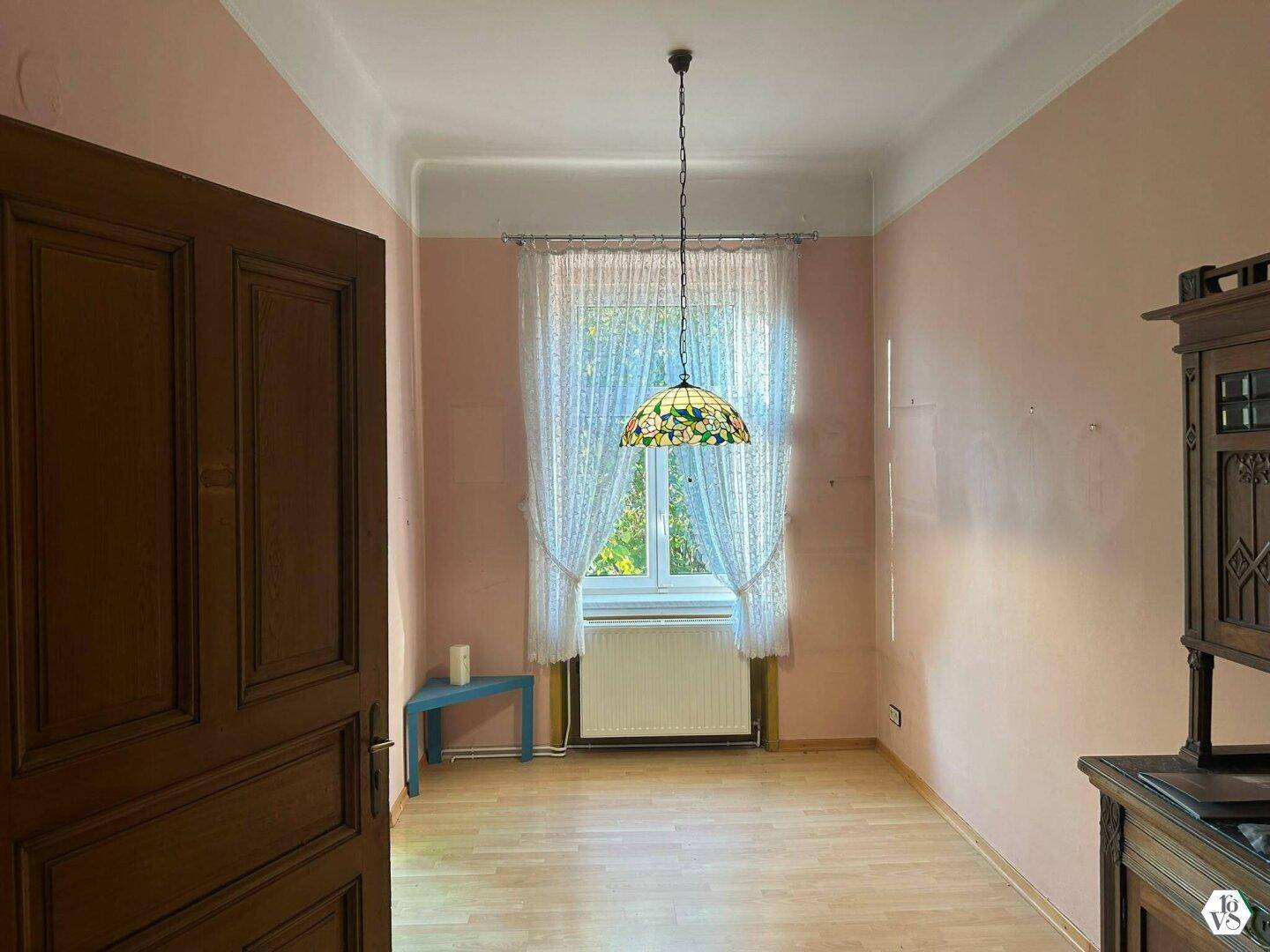 Leeres Zimmer mit rosa Wänden, einer Holztür und einem Fenster mit hellen Vorhängen.