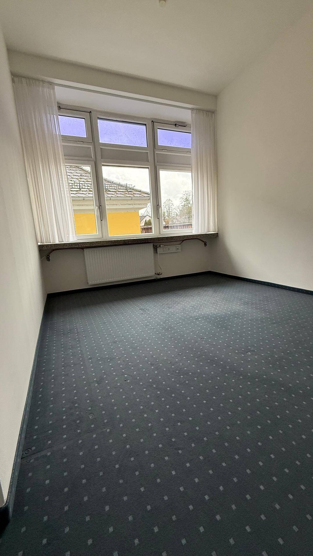 Kleiner, heller Raum mit Fenster, Heizkörper und dunklem Teppichboden, ideal als Arbeitszimmer.