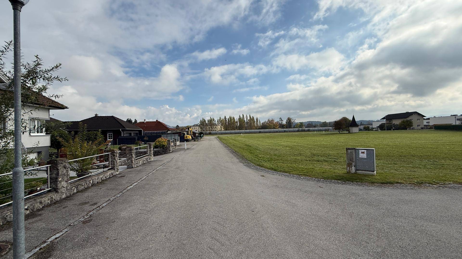 Wohnstraße in ländlicher Umgebung mit Häusern auf der linken Seite und weiten Grünflächen.