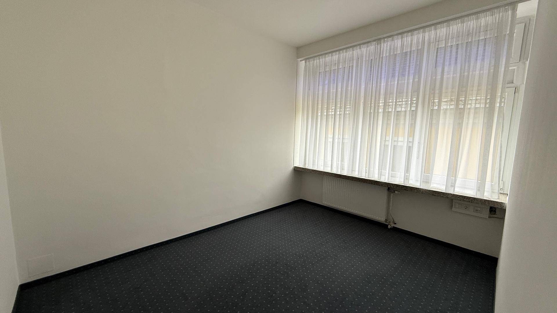 Heller Büroraum mit großem Fenster, Heizkörper und dunklem Teppichboden, bietet viel Tageslicht.