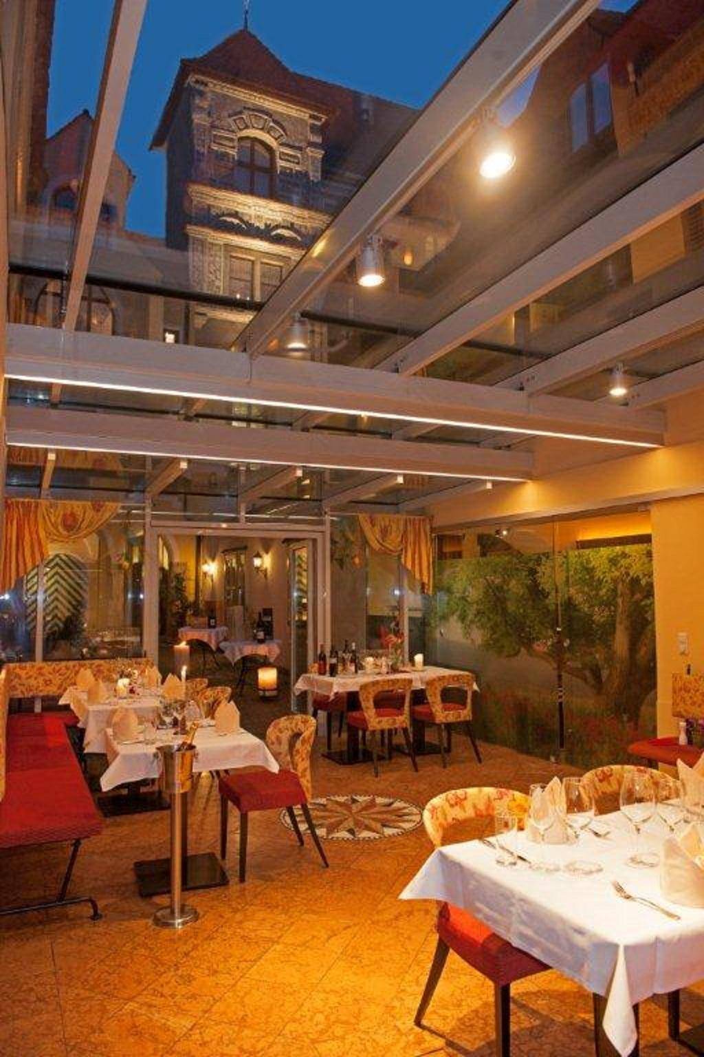Heller Restaurantbereich mit Glasdach, großen Fenstern und festlich gedeckten Tischen, mit Blick auf historische Gebäude.