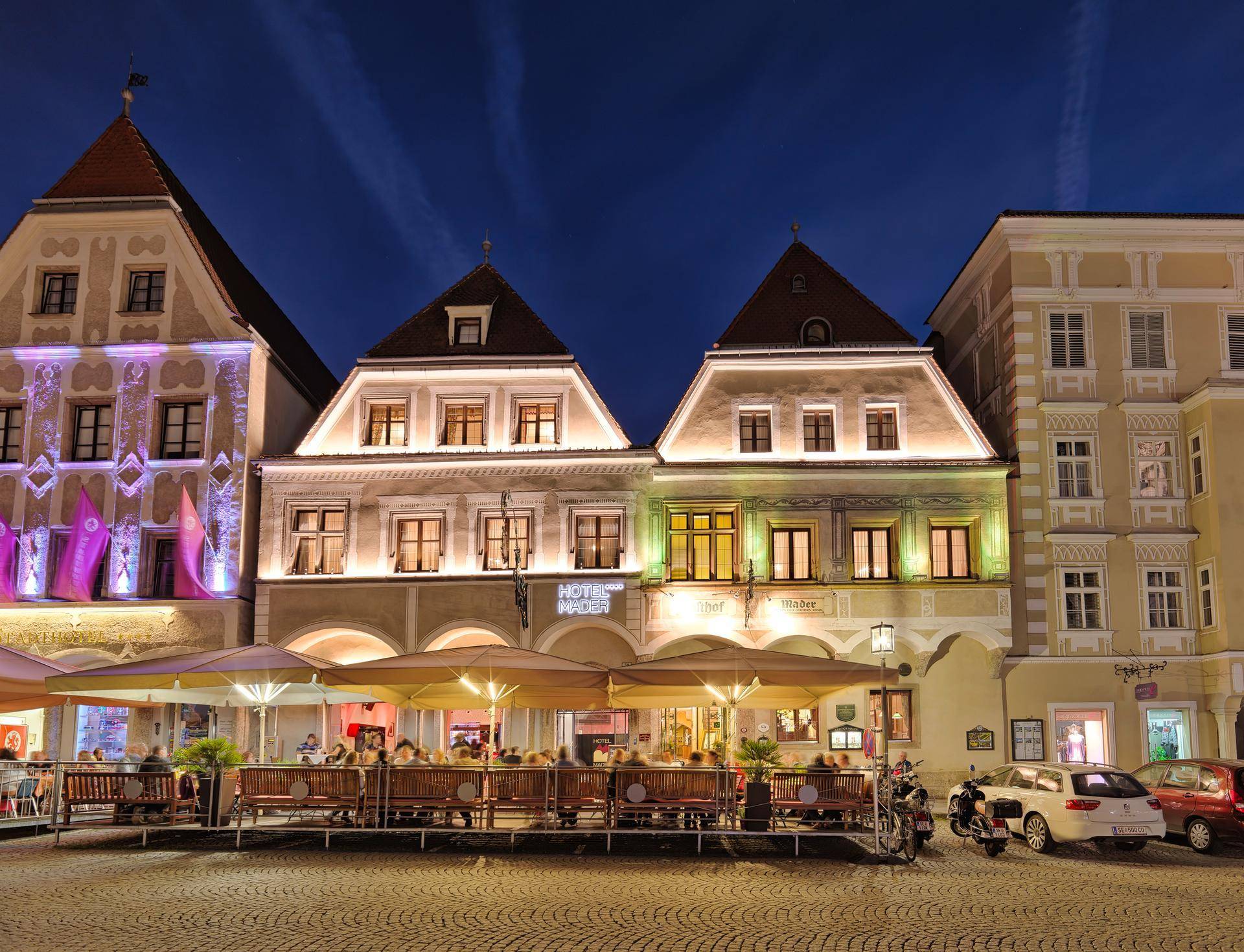 Historische Gebäudefronten am Abend mit beleuchteten Fenstern und einer belebten Außenterrasse.