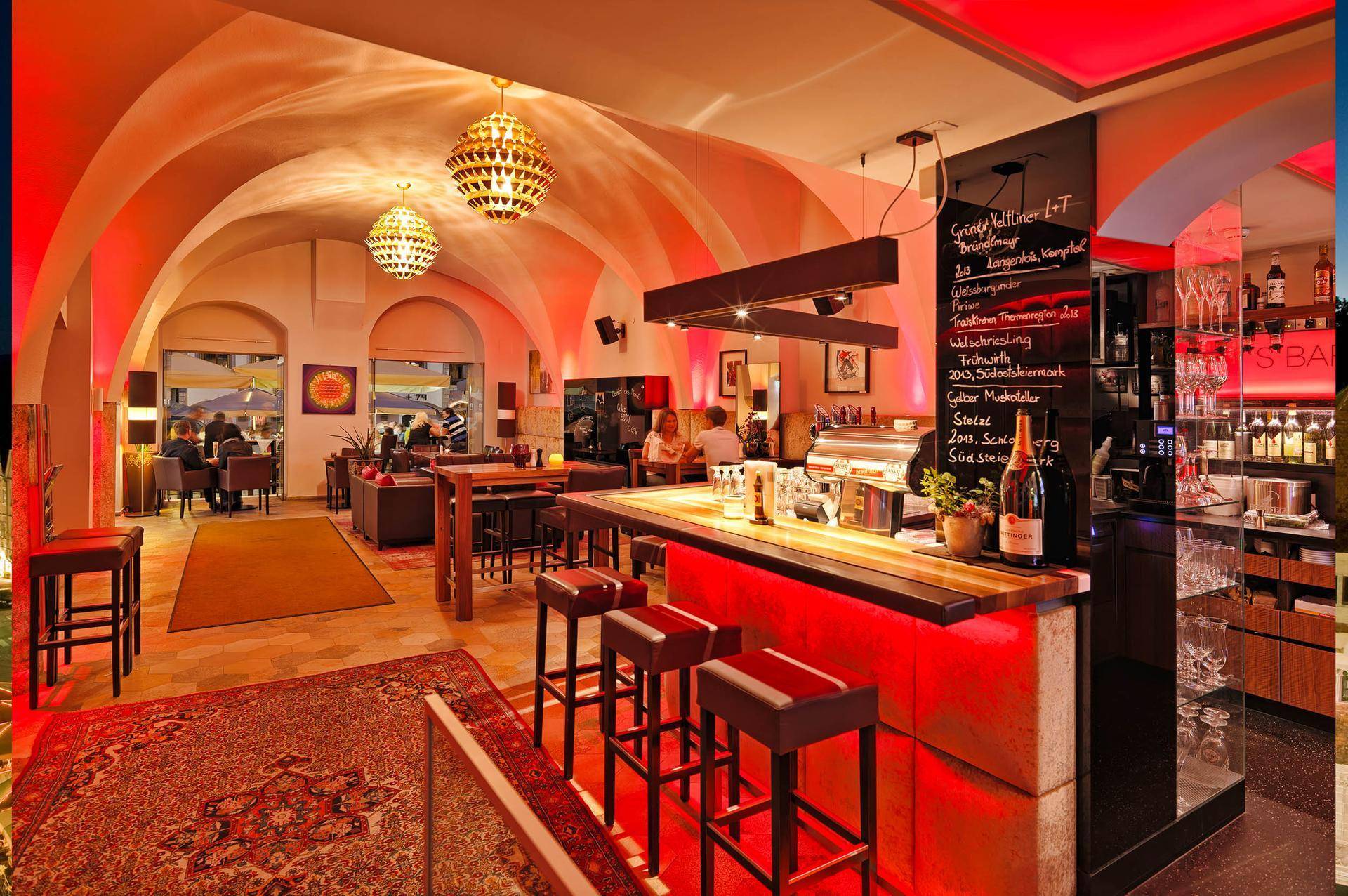 Moderne Bar in einem historischen Gewölbekeller mit roter Beleuchtung, hohen Hockern und gemütlicher Atmosphäre.