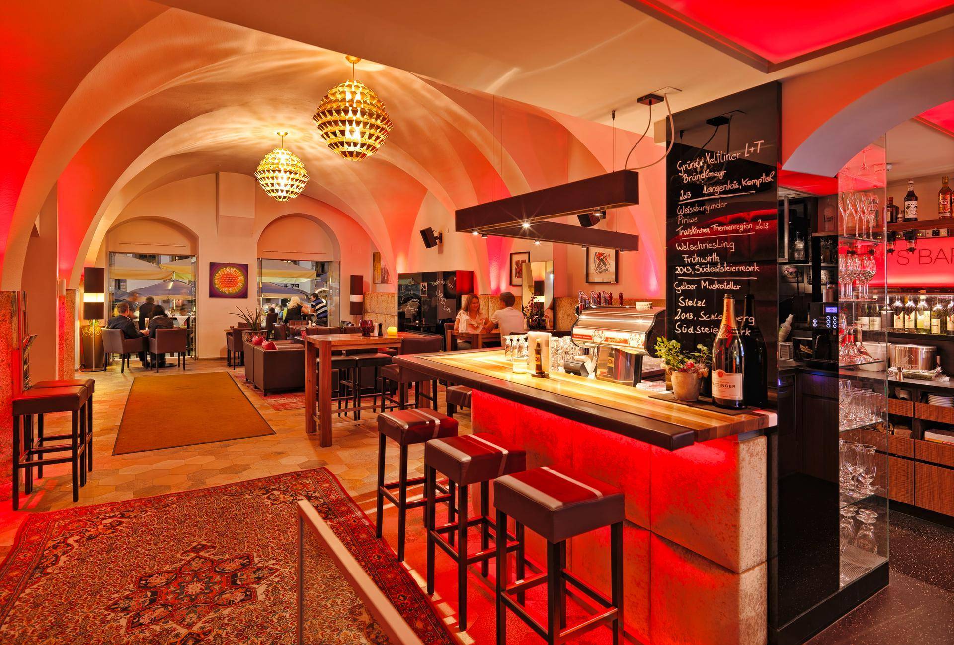 Moderne Bar in einem historischen Gewölbekeller mit roter Beleuchtung, hohen Hockern und gemütlicher Atmosphäre.