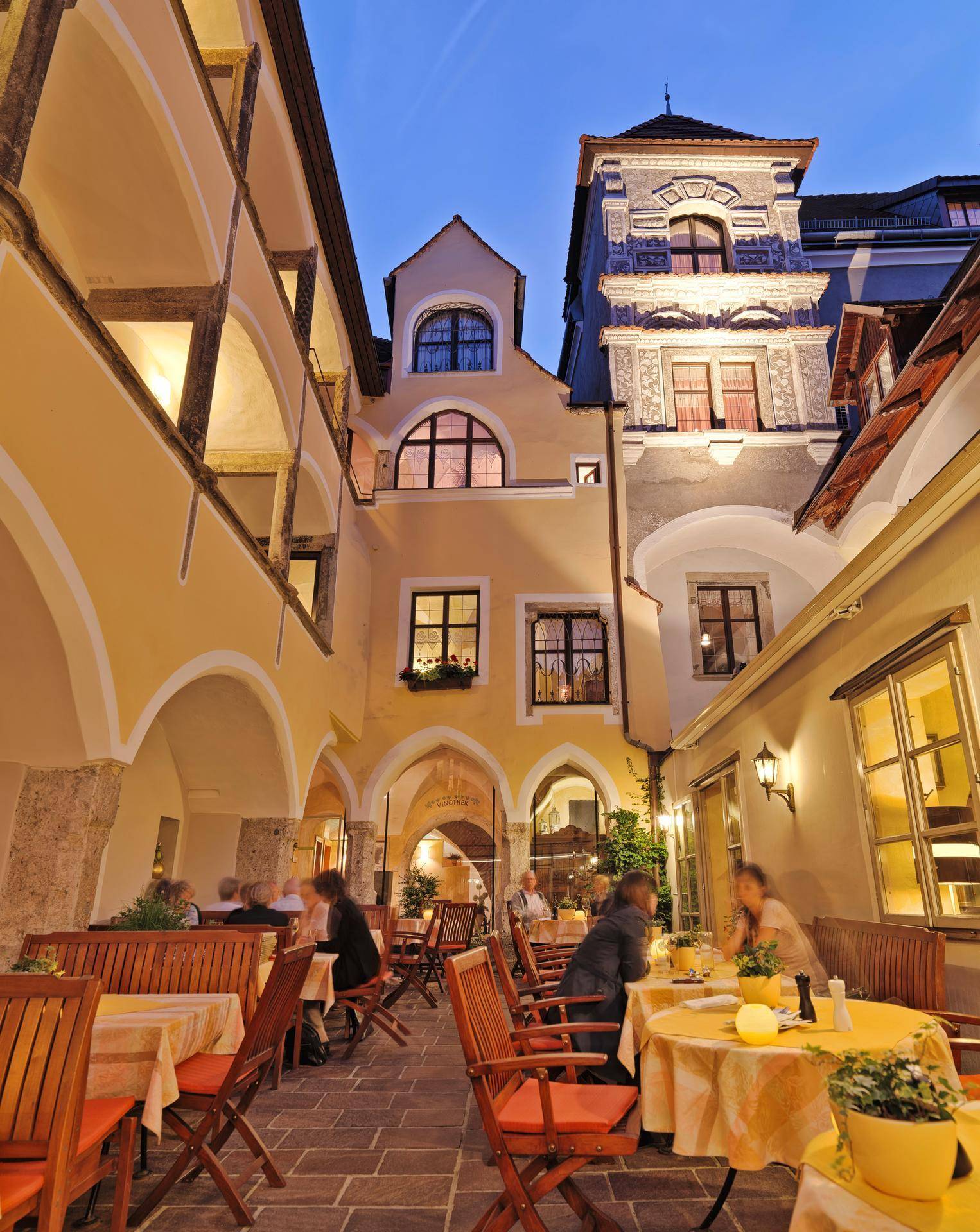 Beleuchteter Innenhof eines historischen Gebäudes am Abend mit Tischen und Stühlen für Gäste.